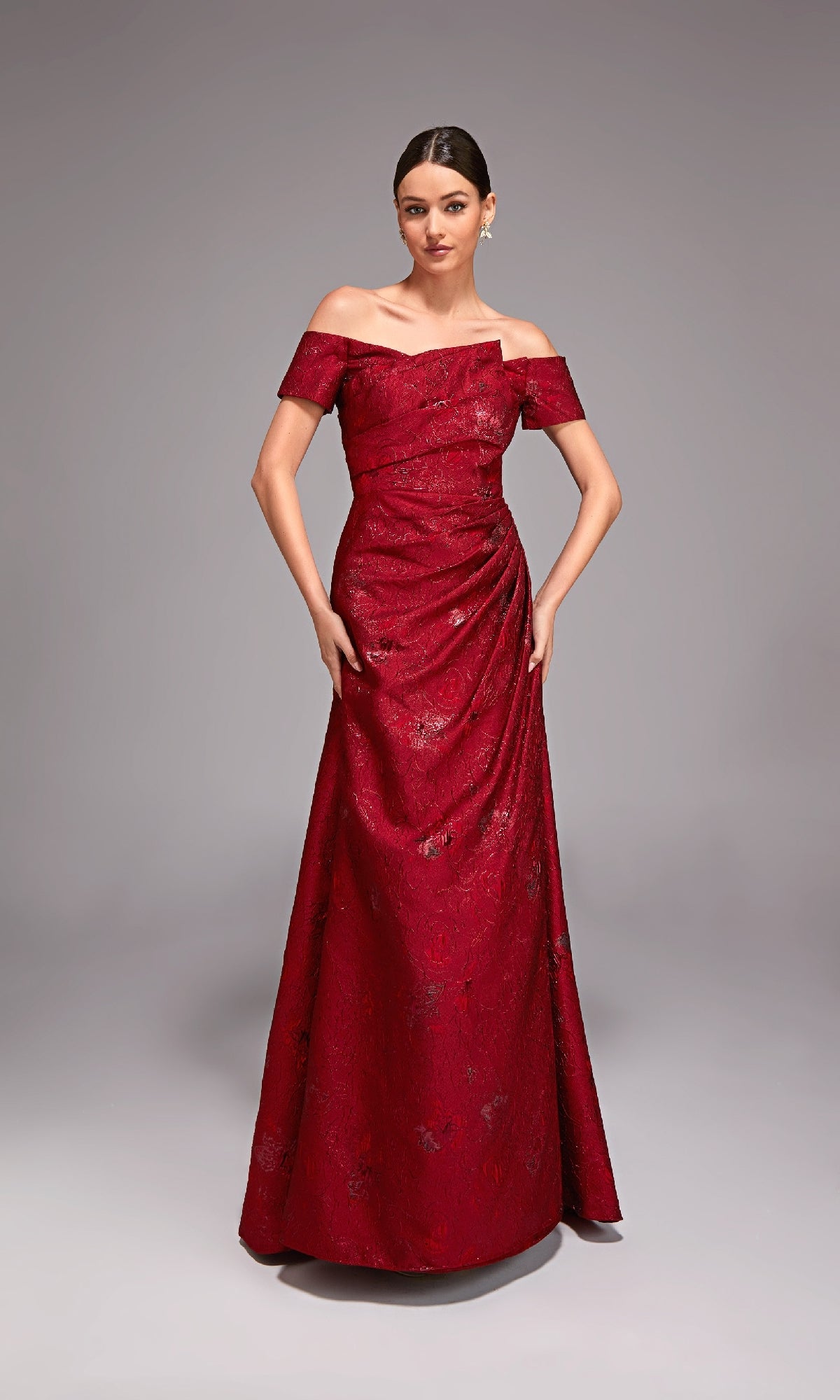 A woman poses in the Alyce 27828 Long Formal Dress—a floor-length, off-the-shoulder red jacquard gown with subtle floral patterns—against a plain gray background.